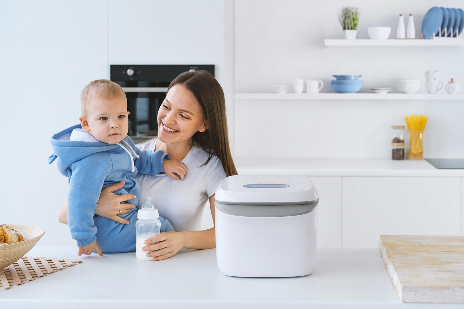 A smiling woman holding a baby in a blue hoodie, sitting at a kitchen counter with a white bread machine and a baby bottle.