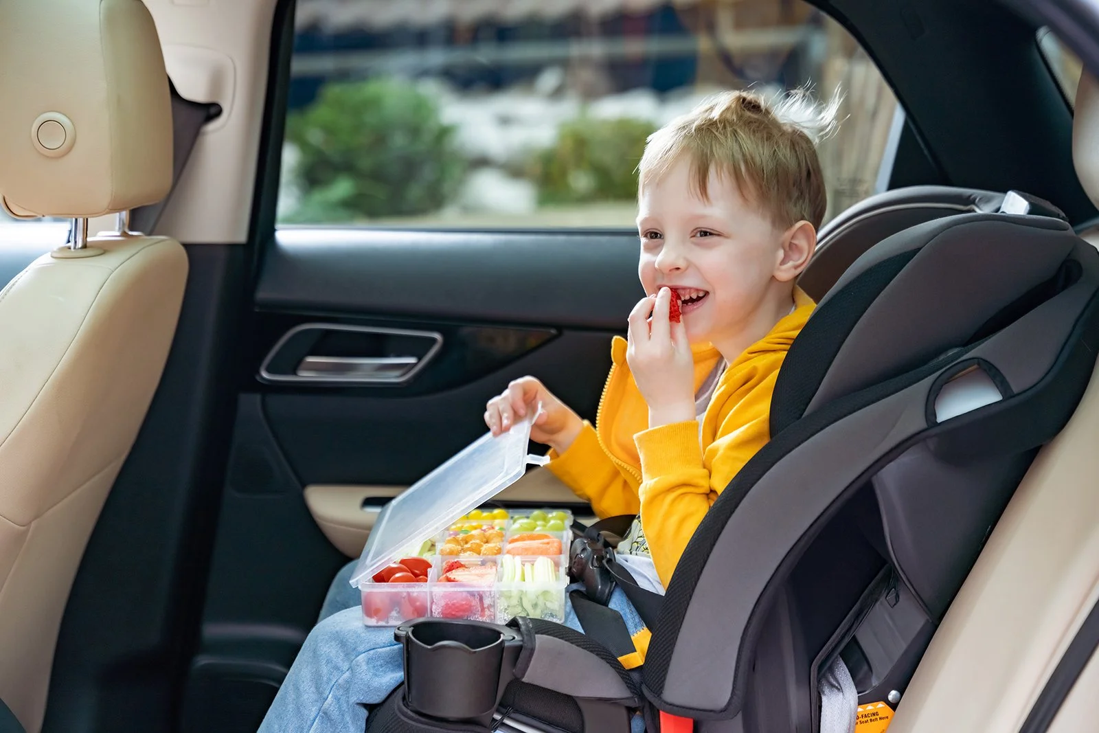 A young boy sitting in a car seat in the back of a vehicle, wearing a yellow jacket, happily eating fruit from a clear plastic container while holding a fruit snack in his hand.