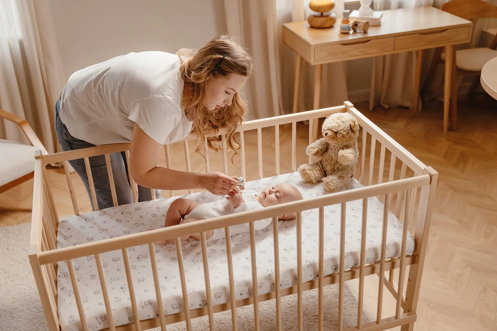 A woman leaning over a crib, feeding a baby with a plush teddy bear sitting inside the crib. The room has wooden floors and furniture, with soft natural lighting.