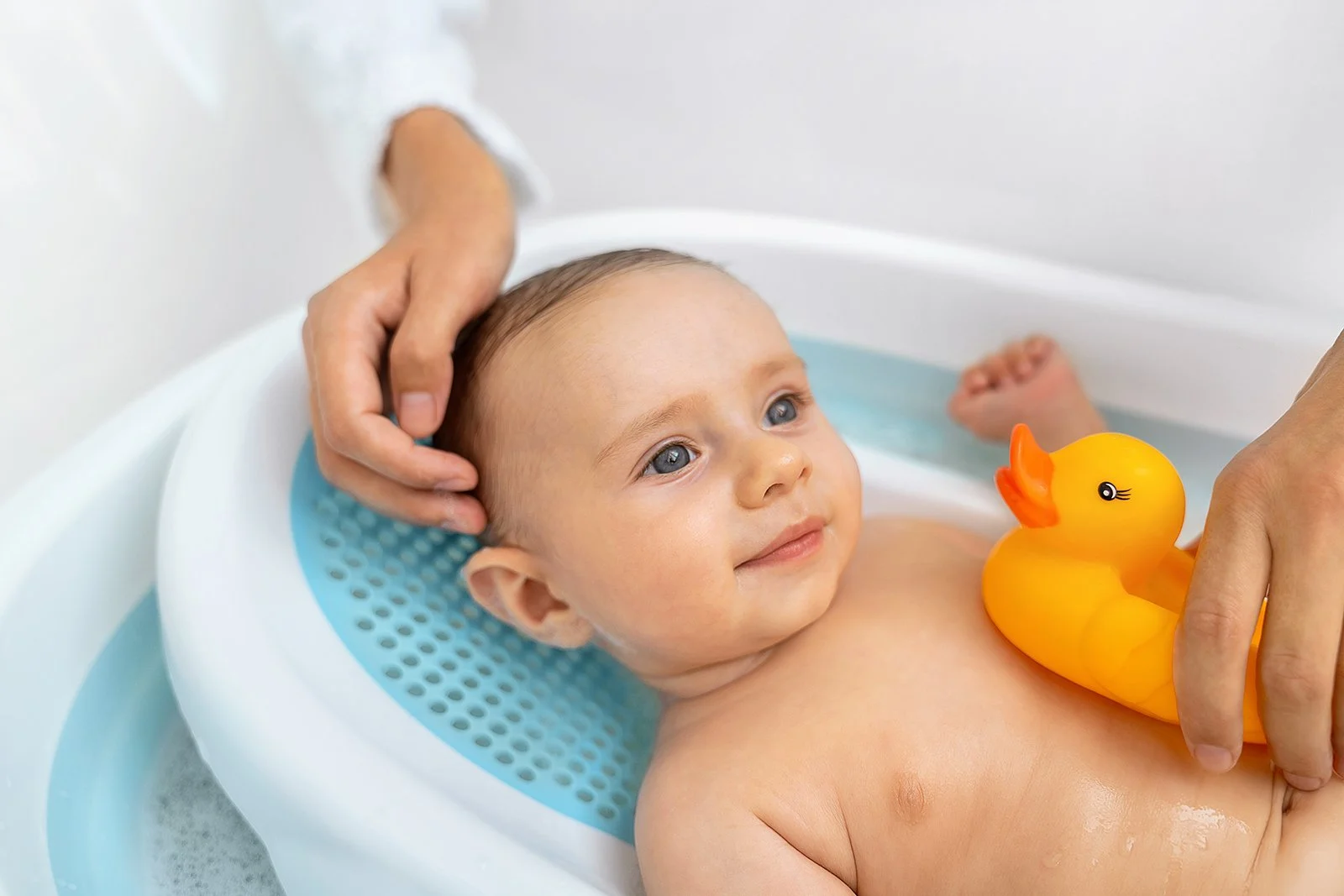 Baby in a bath with a yellow rubber duck, being gently touched by adult hands.