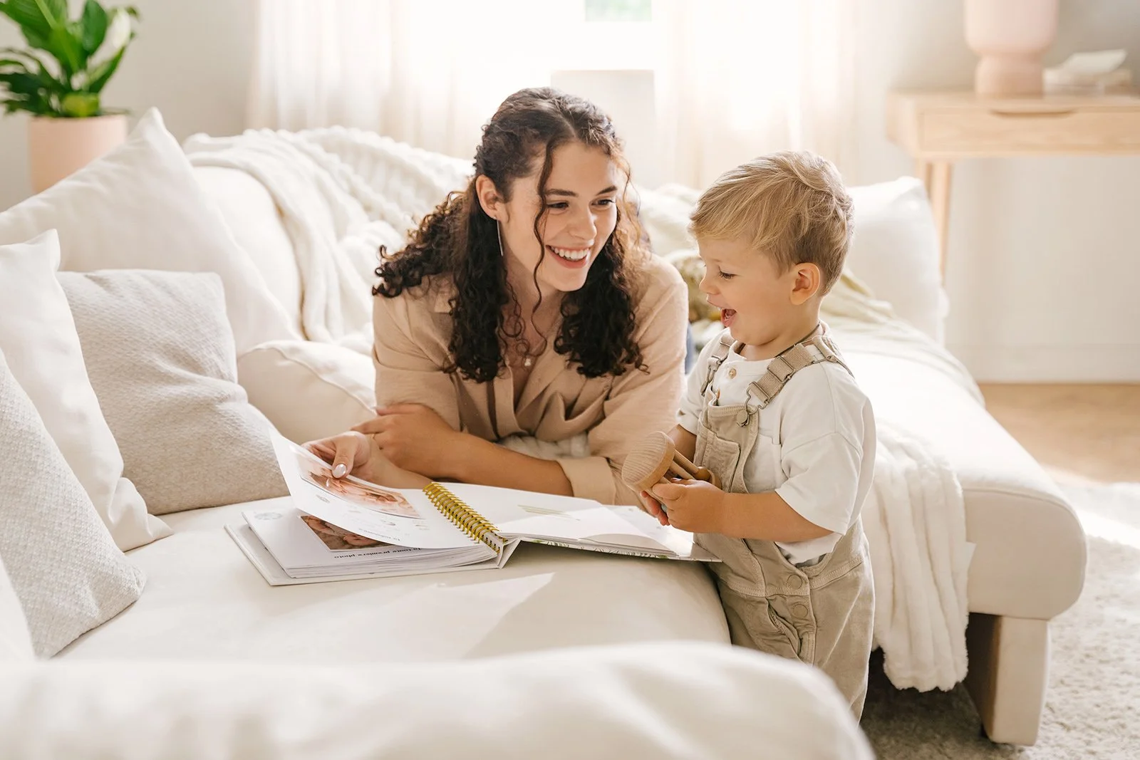 A woman and a young boy sharing a moment on a white sofa in a well-lit living room. The woman is holding a photo album while the boy holds a wooden toy and smiles.