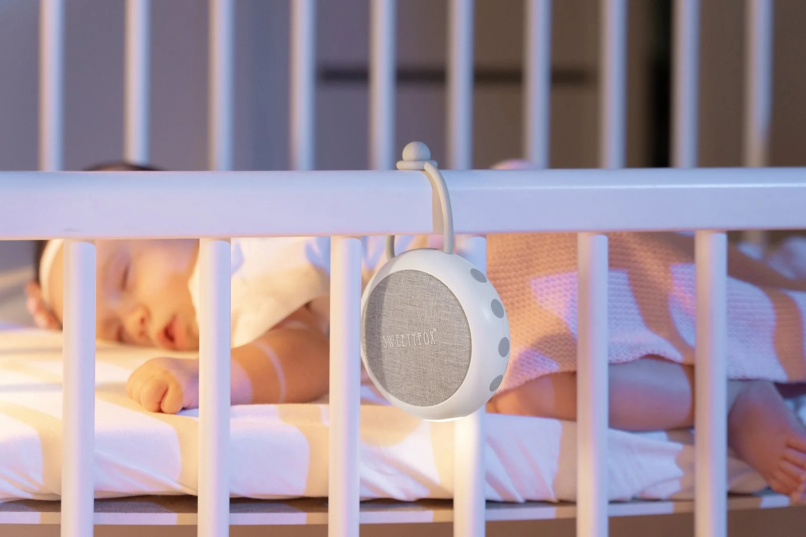 A sleeping baby in a crib with a portable speaker hanging on the rail, showing a cozy nursery scene.