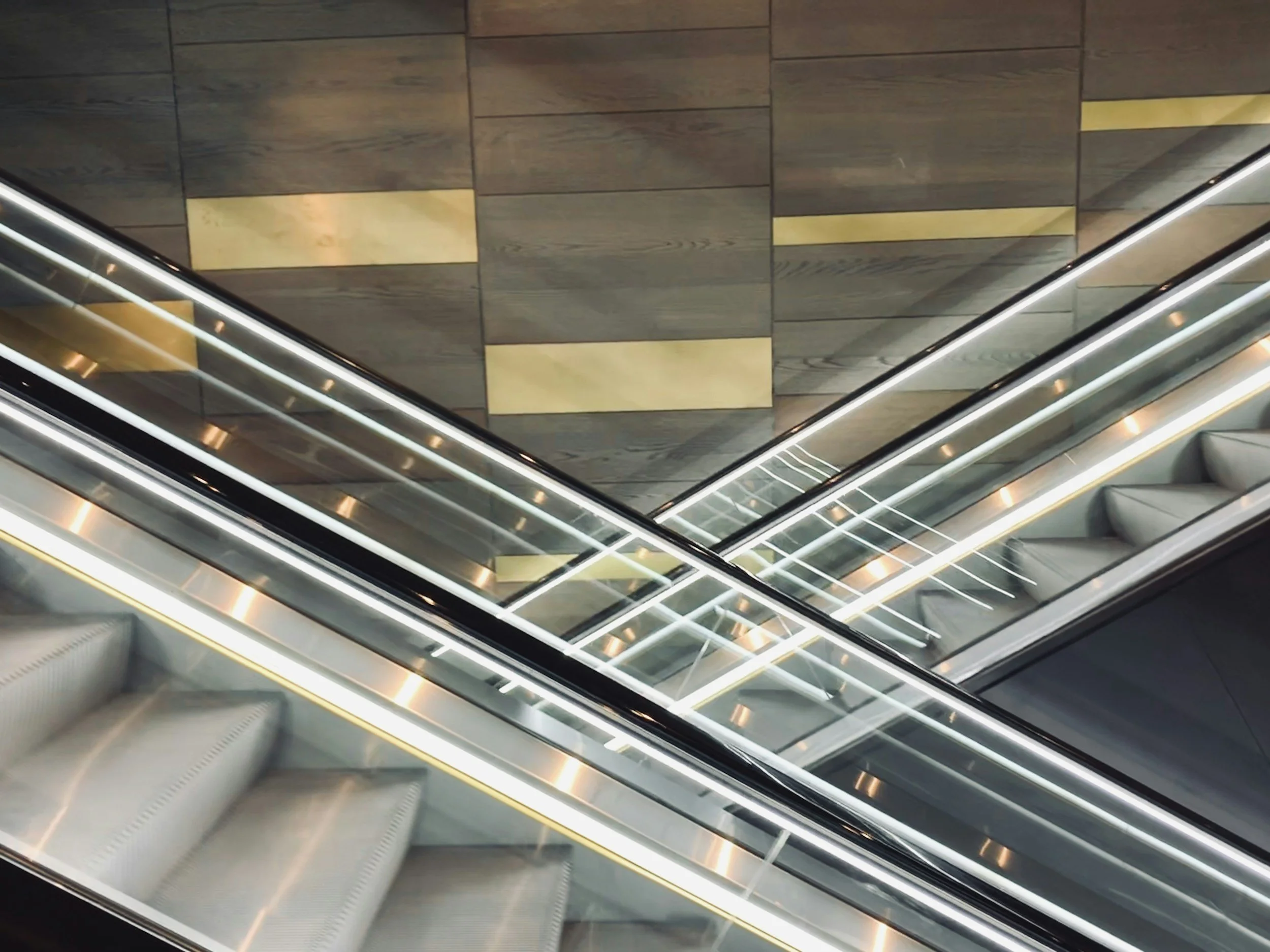 Top-down view of a modern, illuminated escalator in a building with wooden and gold accents on the floor.