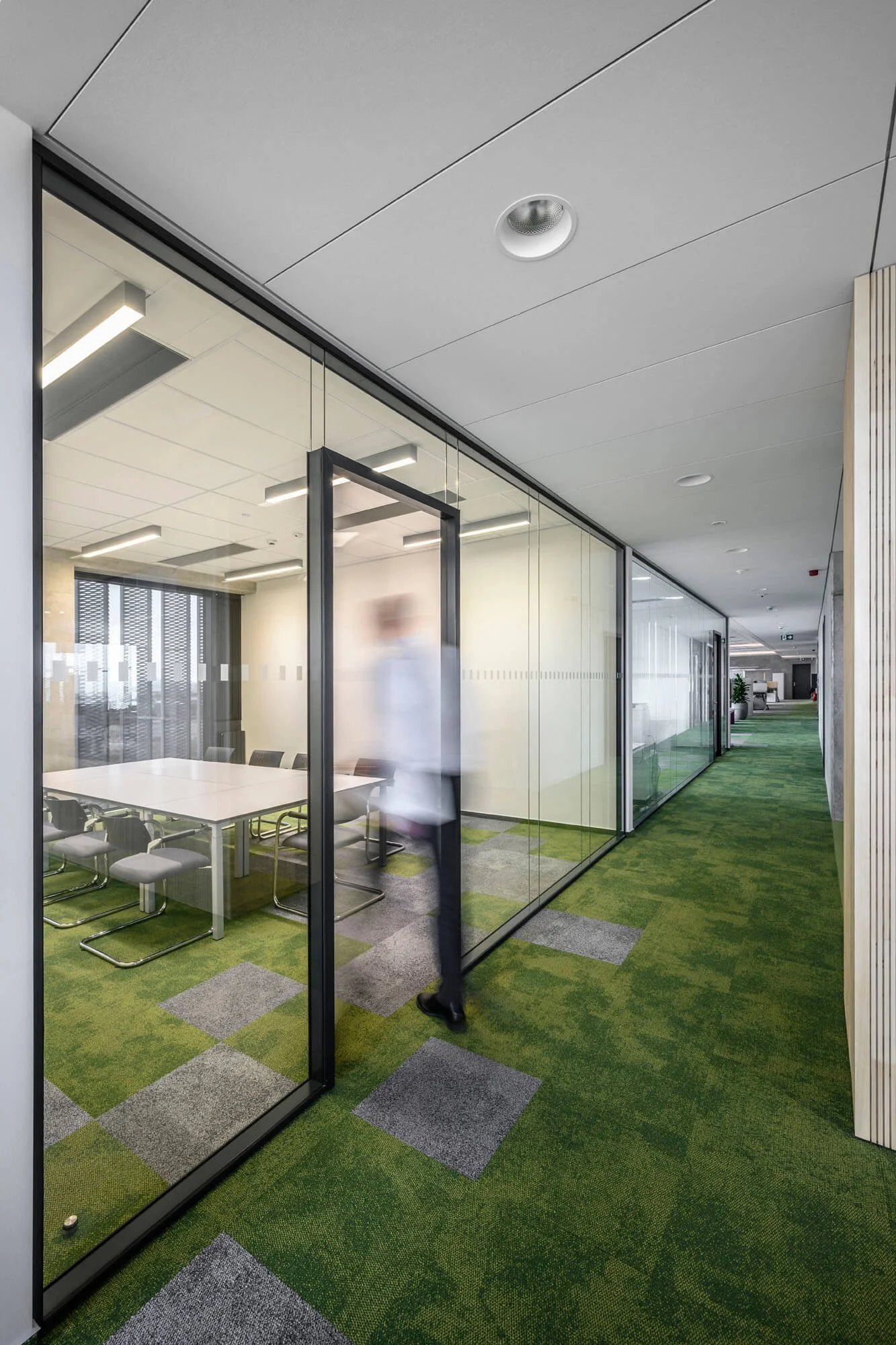Büroflur mit Glaswänden, grüner Teppich, moderne Deckenbeleuchtung und ein sitzungsraum im Hintergrund, eine Person geht vorbei.