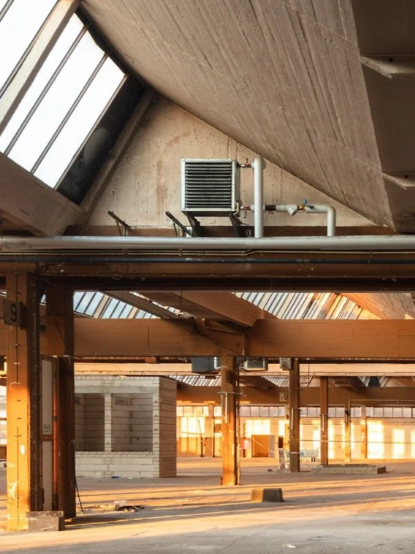 Leere Industriehalle mit Sonnenlicht, sichtbare Dachstrukturen, Lüftungsanlage und mehreren Stützpfeilern.