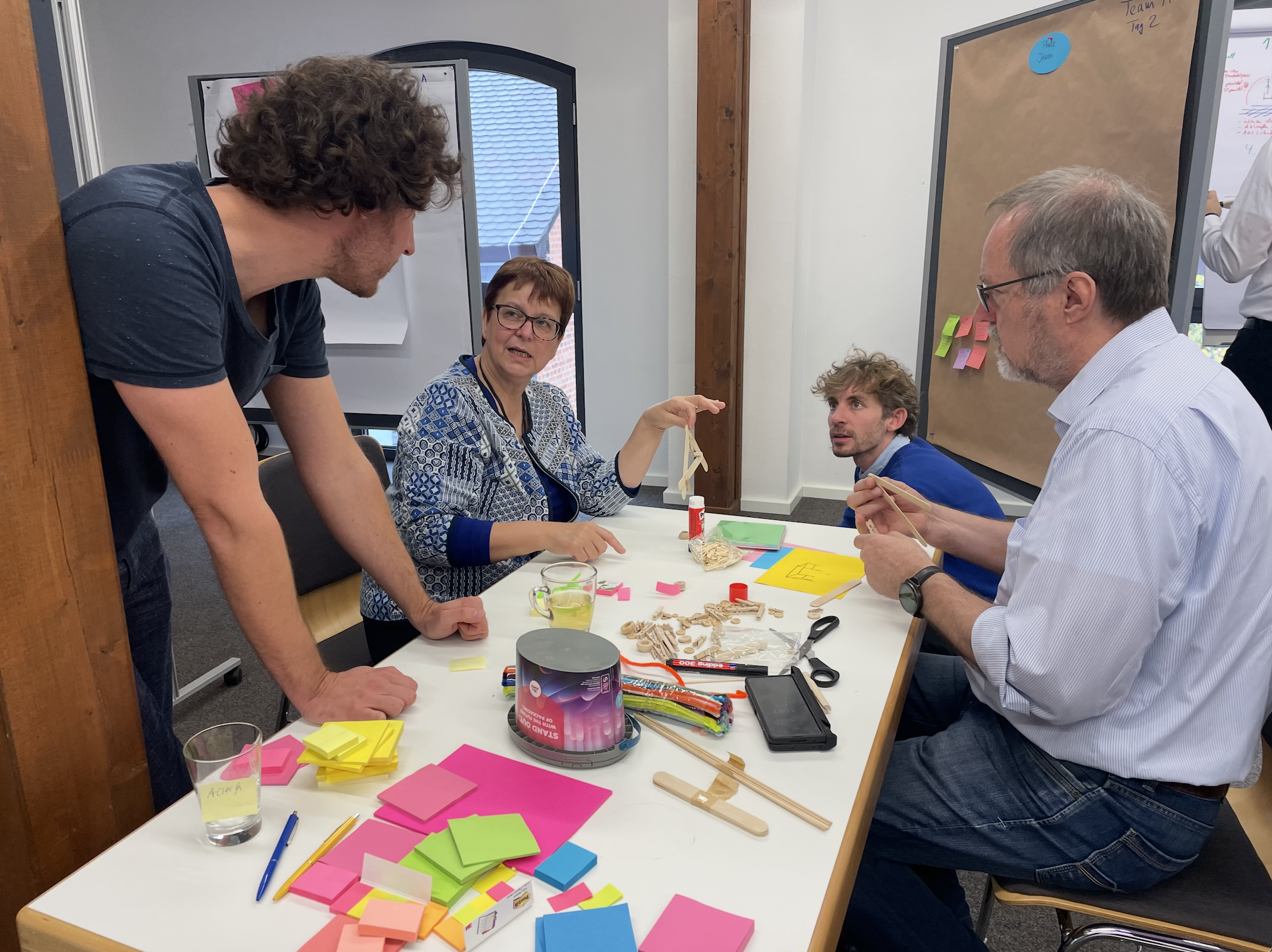 Vier Personen arbeiten bei einem Workshop an einem Tisch mit bunten Post-its, Bastelmateria, Schere und Stiften. One woman erklärt etwas den anderen, während zwei Männer zuhören.