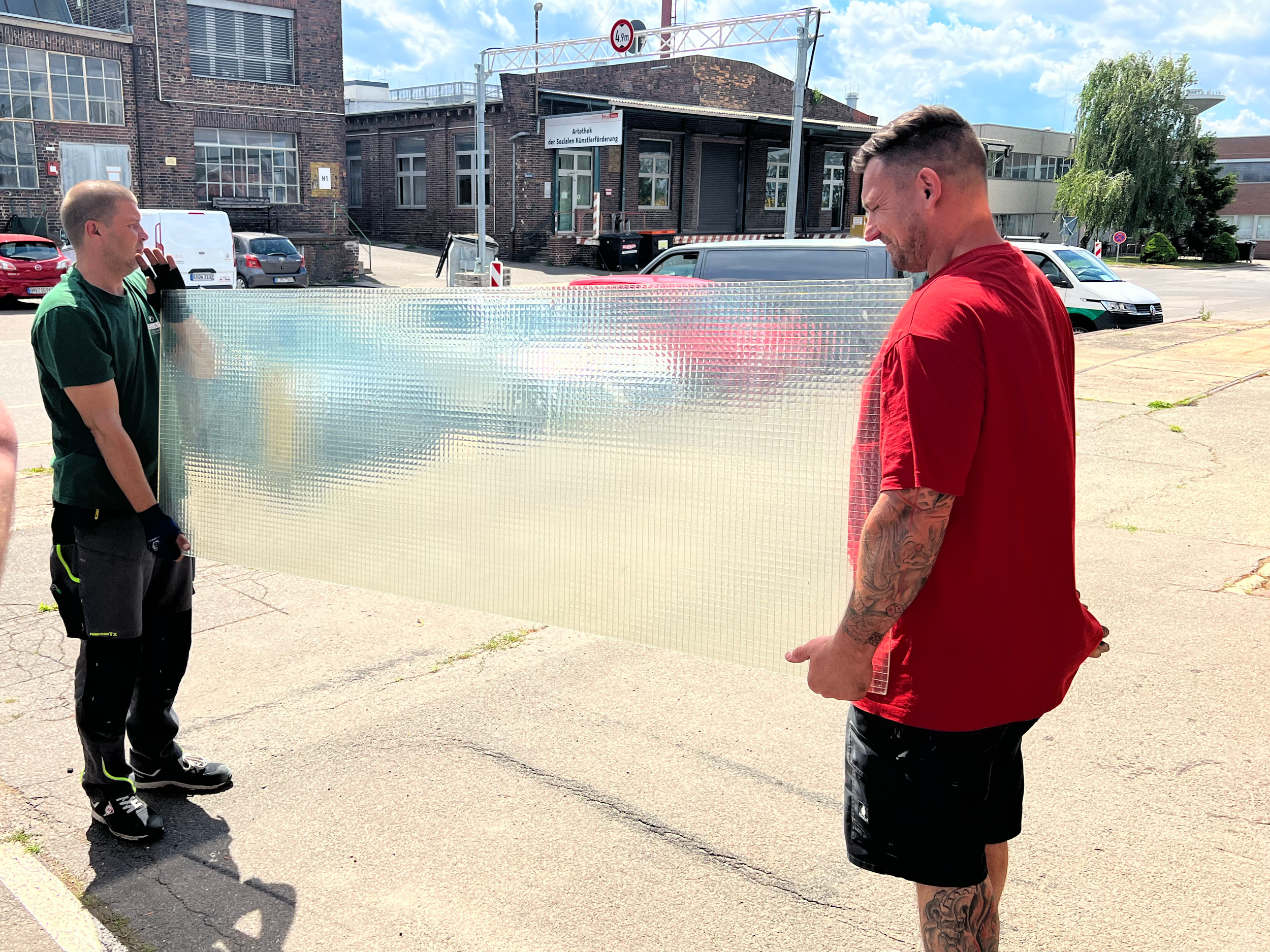 Zwei Männer stehen im Freien, einer hält eine große, transparente, bemusterte Acrylplatte. Der Mann auf der linken trägt ein grünes T-Shirt und schwarze Arbeitshosen, der rechte trägt ein rotes T-Shirt und schwarze Shorts, beide sind lachend.