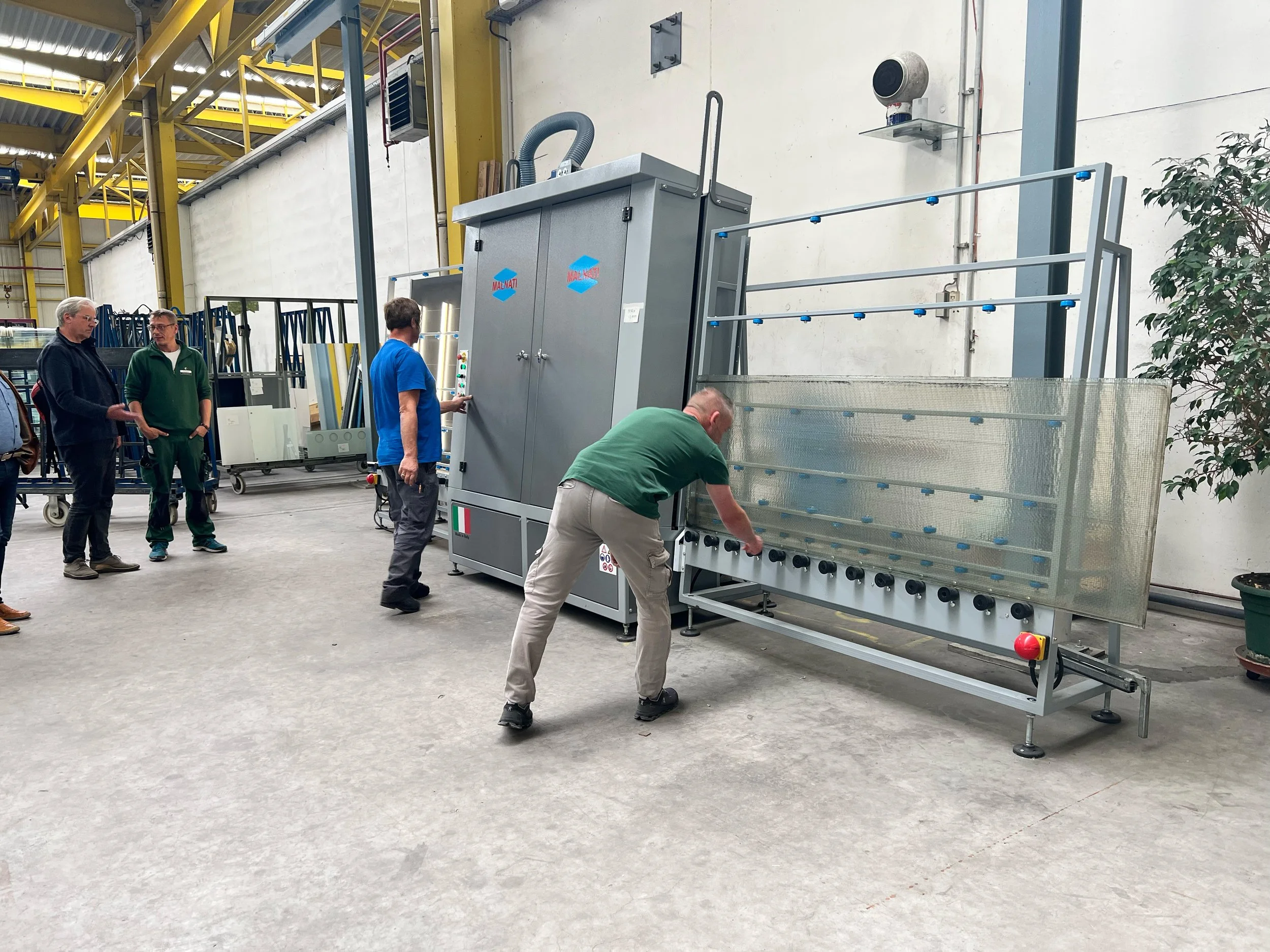 Mehrere Personen in einer Fabrikhalle arbeiten mit einer Maschine, einige beobachten, einer bedient die Maschine, andere stehen Schlange. Die Maschine ist groß und grau mit blauen Markierungen.