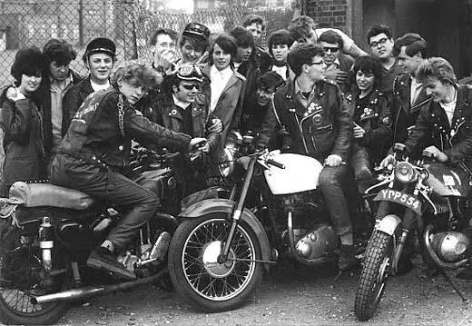 Group of young people, mostly teenagers, gathered around two vintage motorcycles on a street. Some wear leather jackets and sunglasses, with one person sitting on a motorcycle. The background shows a chain-link fence and brick buildings.