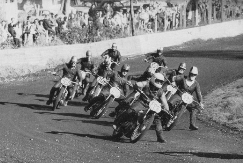 Vintage motorcycle race with multiple racers leaning into a turn on a dirt track, spectators watching from behind a fence in the background.