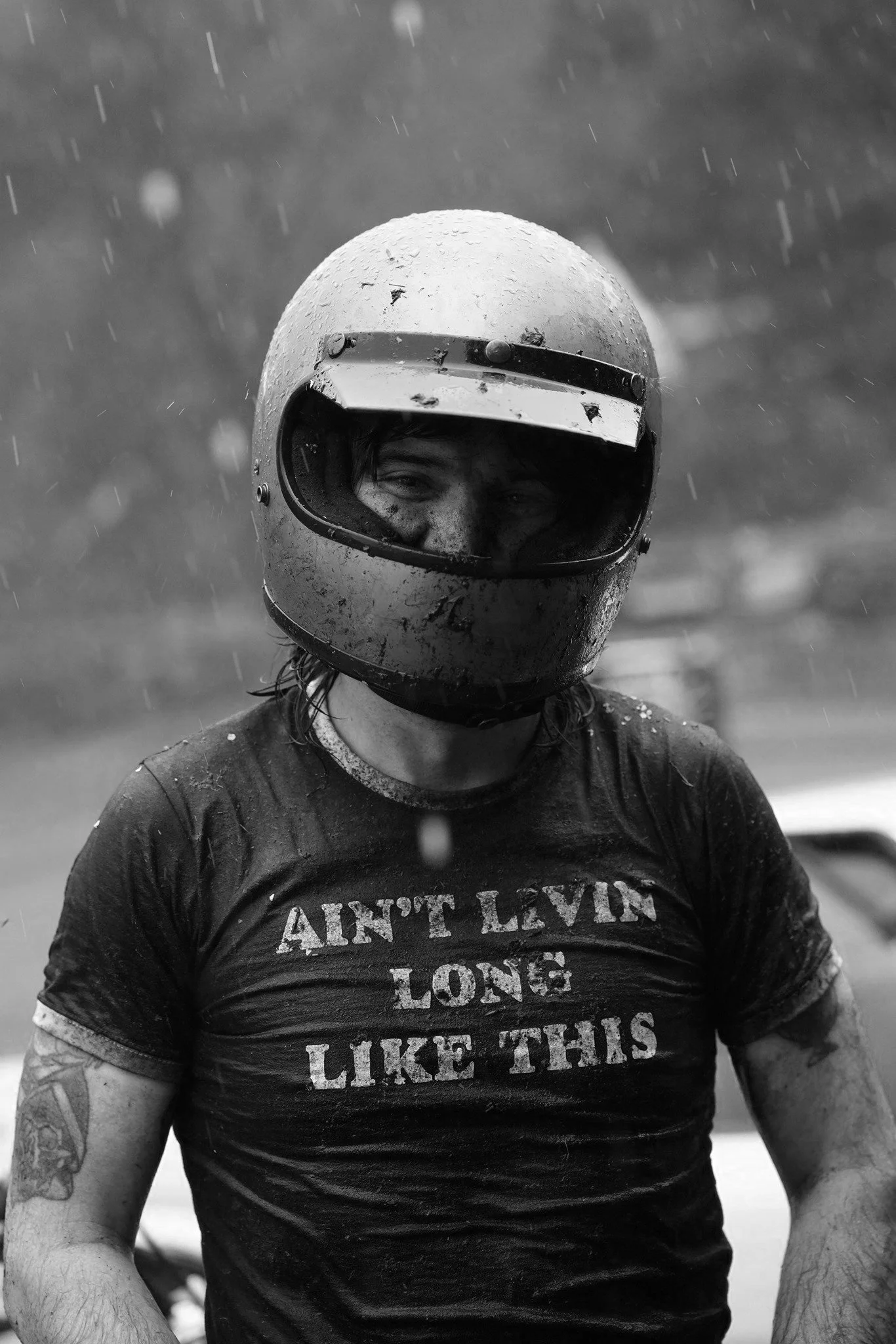 Person wearing a motorcycle helmet and a wet t-shirt with the text "Ain't Leave Long Like This," standing in the rain.