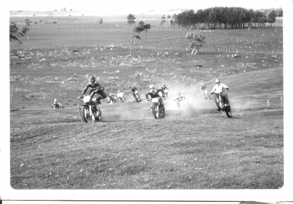 Multiple people riding vintage dirt bikes on a grassy hillside, with more riders in the background and a tree-covered hill in the distance.