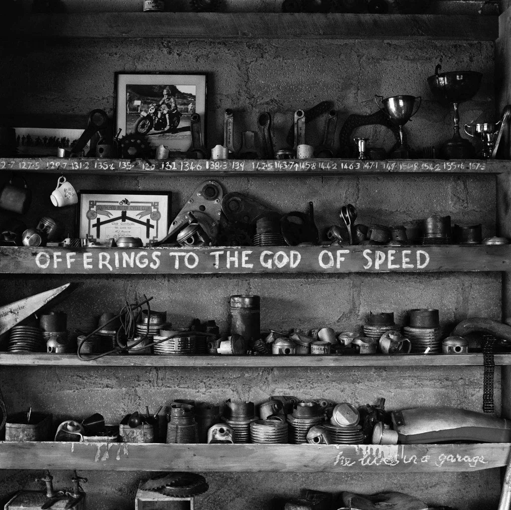 A black and white photograph of a workshop shelf filled with various metal objects, tools, and trophies. A framed photo of a person on a motorcycle and a framed image with a timeline and bicycle are also visible. A hand-painted sign on the shelf reads, 'OFFERINGS TO THE GOD OF SPEED'.