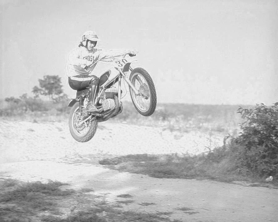 A person wearing a helmet and shirt riding a motorcycle and jumping over a dirt road with bushes and trees in the background.
