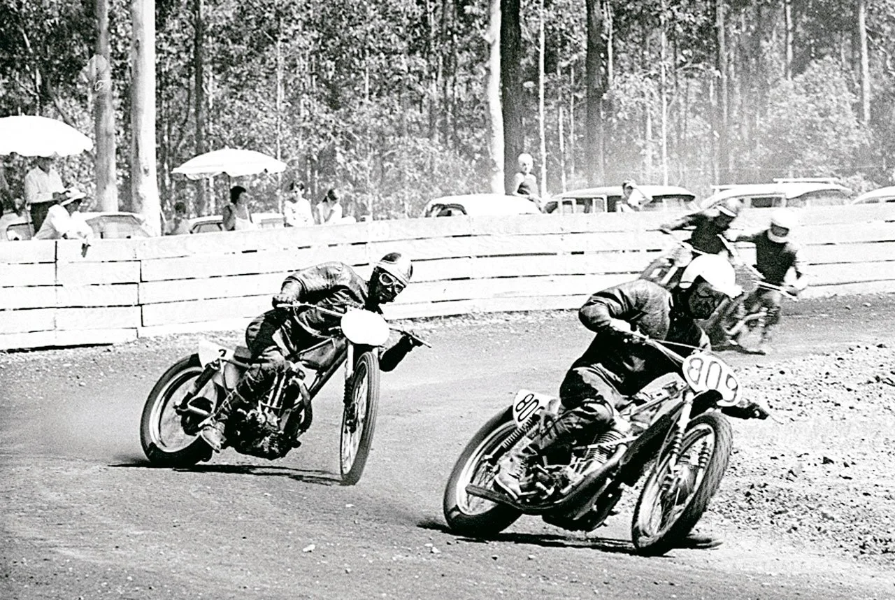 Black and white photo of vintage motorcycle race on a dirt track with three racers leaning into a turn, spectators watching behind a wooden barrier, with cars and trees in the background.