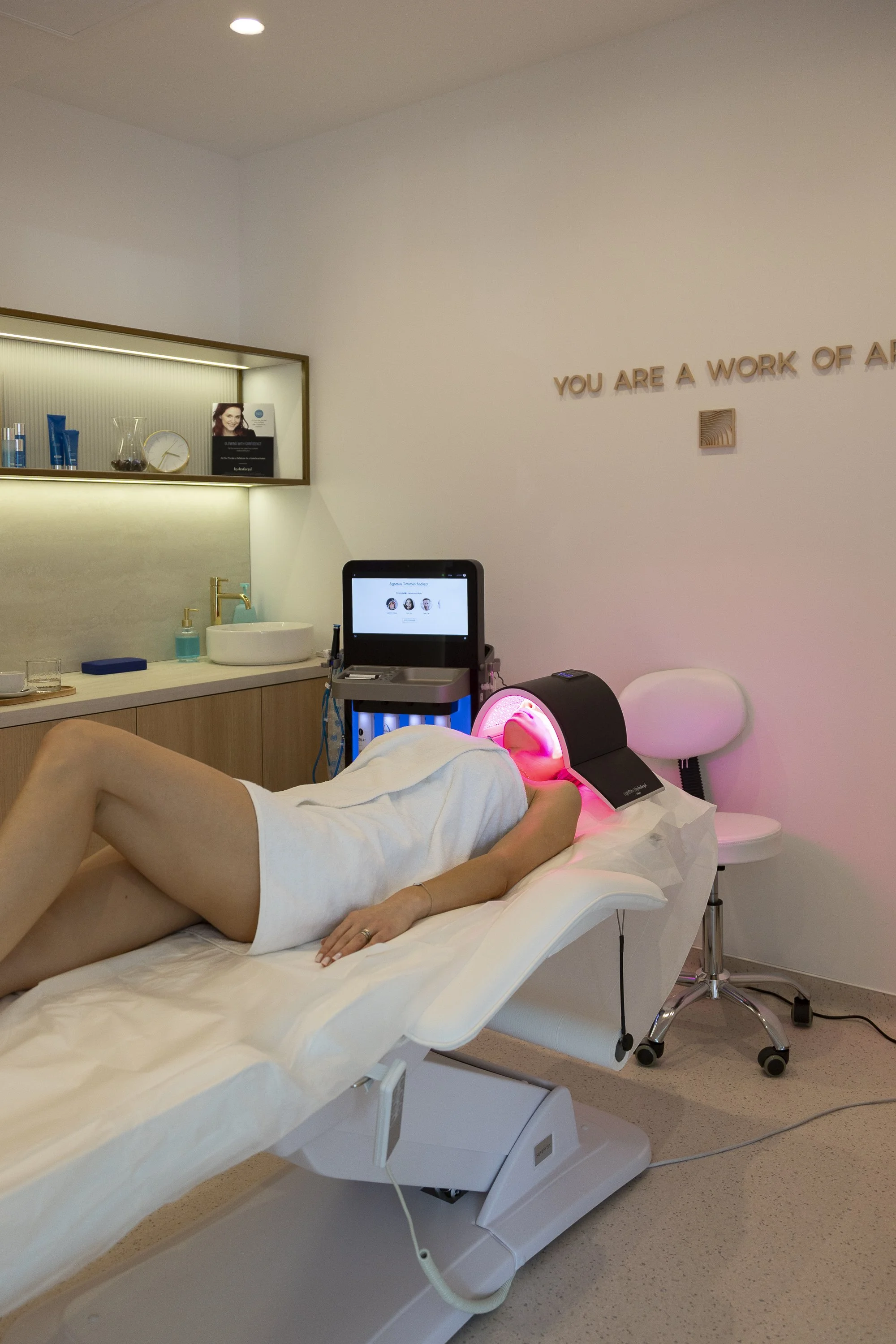 A woman receiving a facial treatment with a pink light therapy device in a spa or clinic room. The room has a beige and white color scheme with a sign on the wall that reads "You are a work of art."