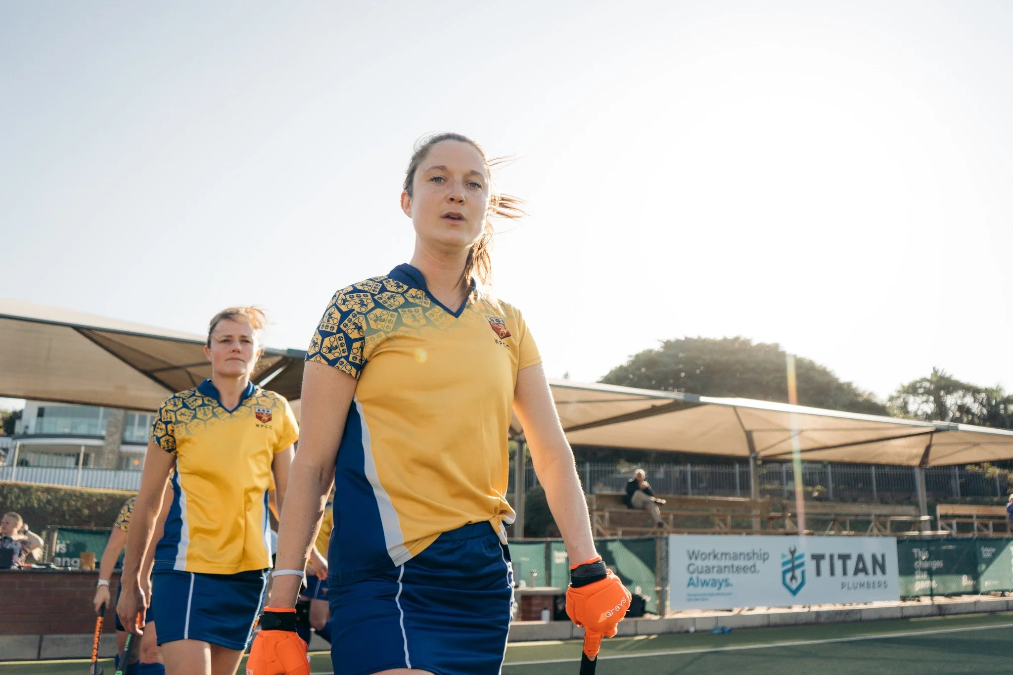 Two female field hockey players in yellow and blue uniforms walking on the field after a game, with sunlight and a shaded seating area in the background.