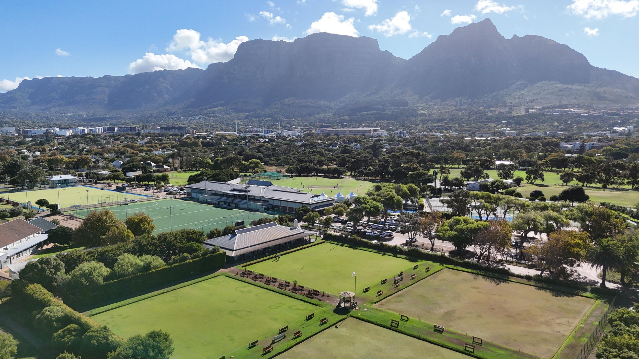 Aerial view of WPCC with tennis courts, cricket fields, and trees, with mountains in the background under a partly cloudy sky.