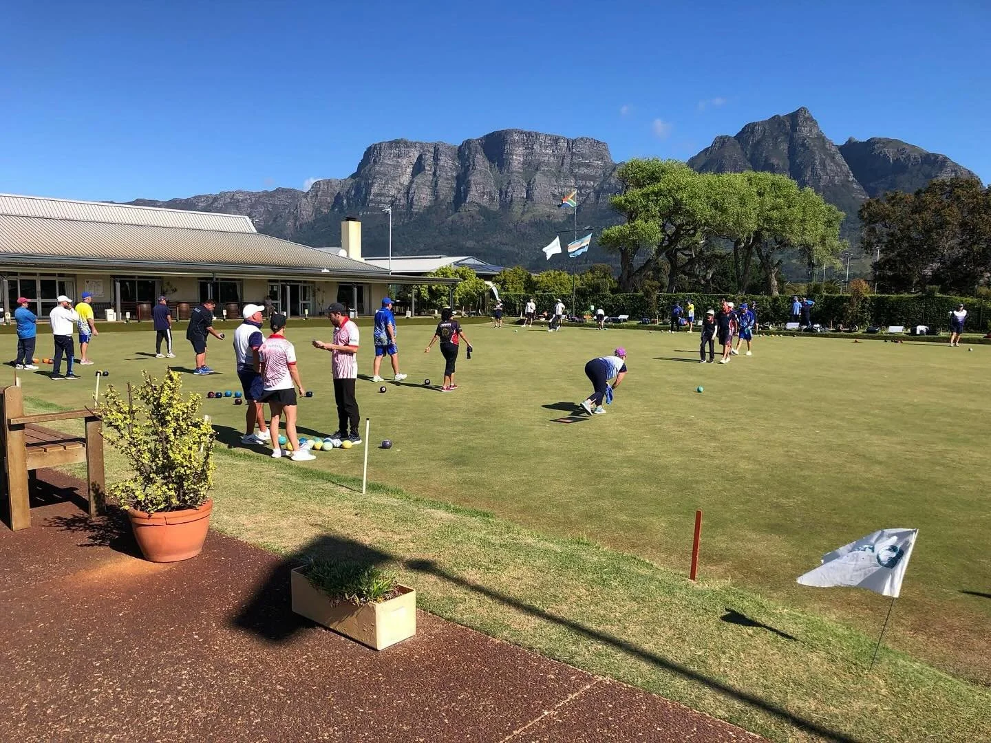 People playing lawn bowls on a green field with a mountain backdrop, trees, and a building in the background under a blue sky.
