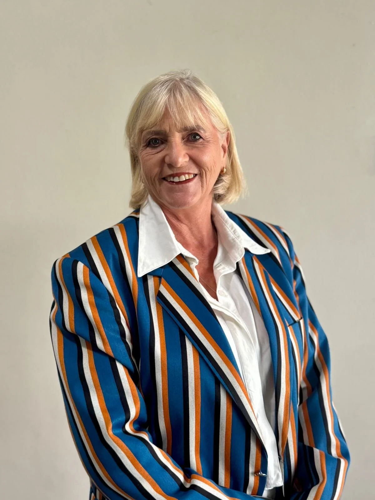 An older woman with blonde hair smiling, wearing a multicolored striped blazer over a white collared shirt, standing against a plain beige wall.