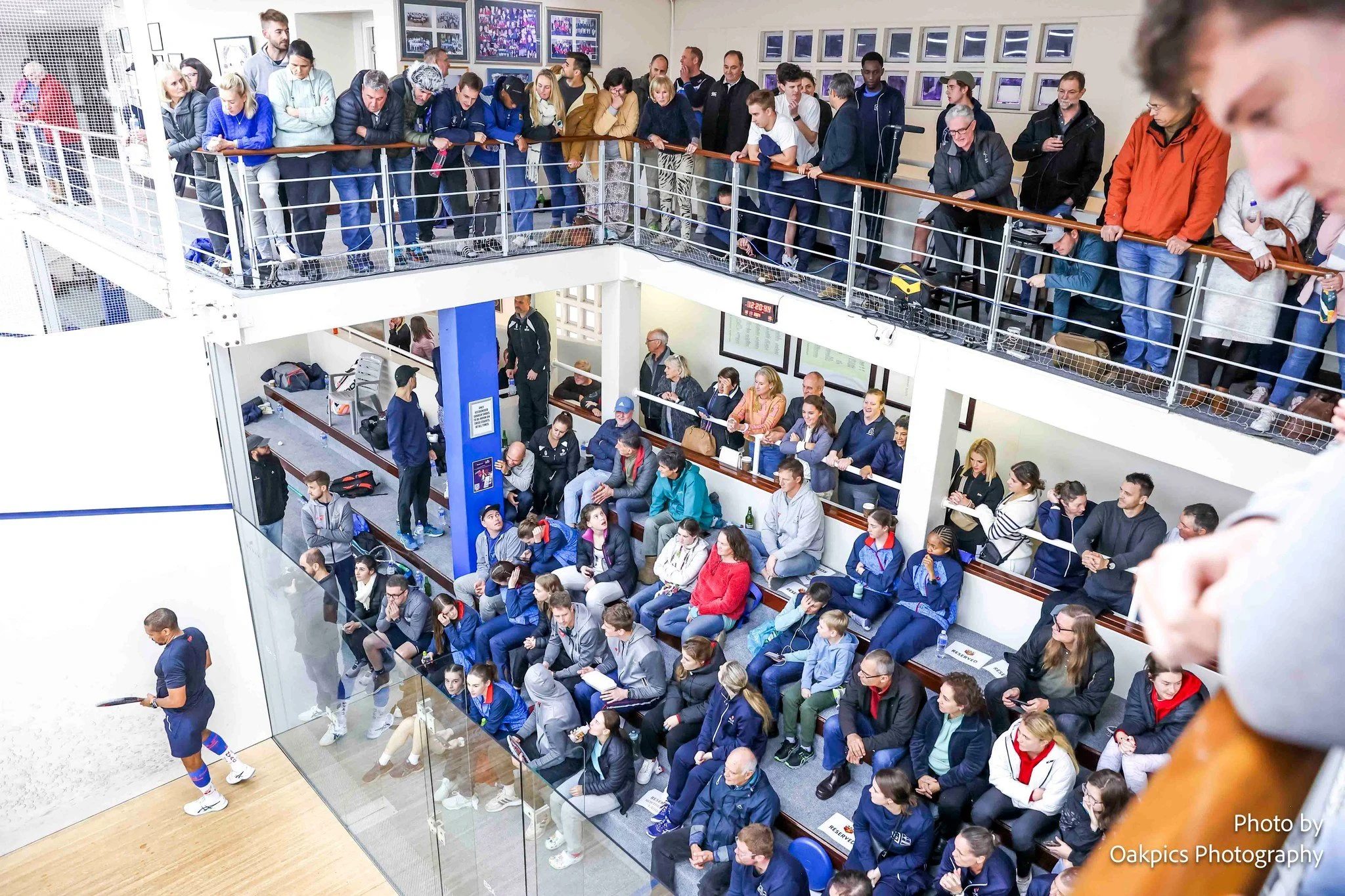 Indoor sports arena with spectators seated in the lower and upper tiers, watching a match. People are wearing casual and sports attire, some seated and others standing, engaged in conversation or observing. A referee or coach stands near the court, which has a glass boundary and a wooden floor.