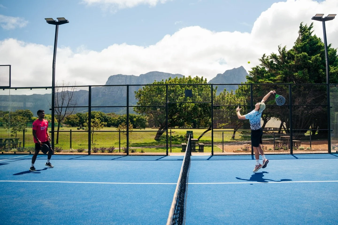 Two men playing pickleball on a blue court outdoors, with mountains and trees in the background. One man in a pink shirt holds a paddle, while the other in a blue patterned shirt jumps to hit a pickleball.