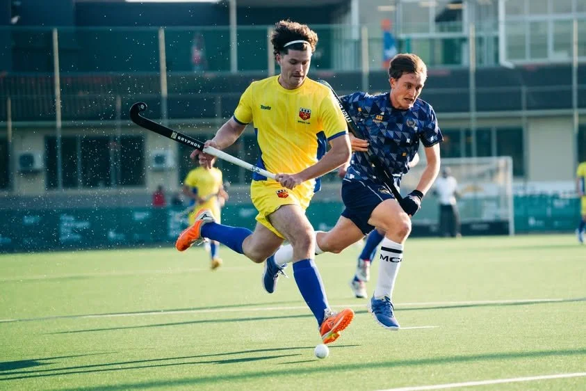 Two male field hockey players competing for the ball on a grass field, with one in a yellow uniform and the other in a dark blue uniform.
