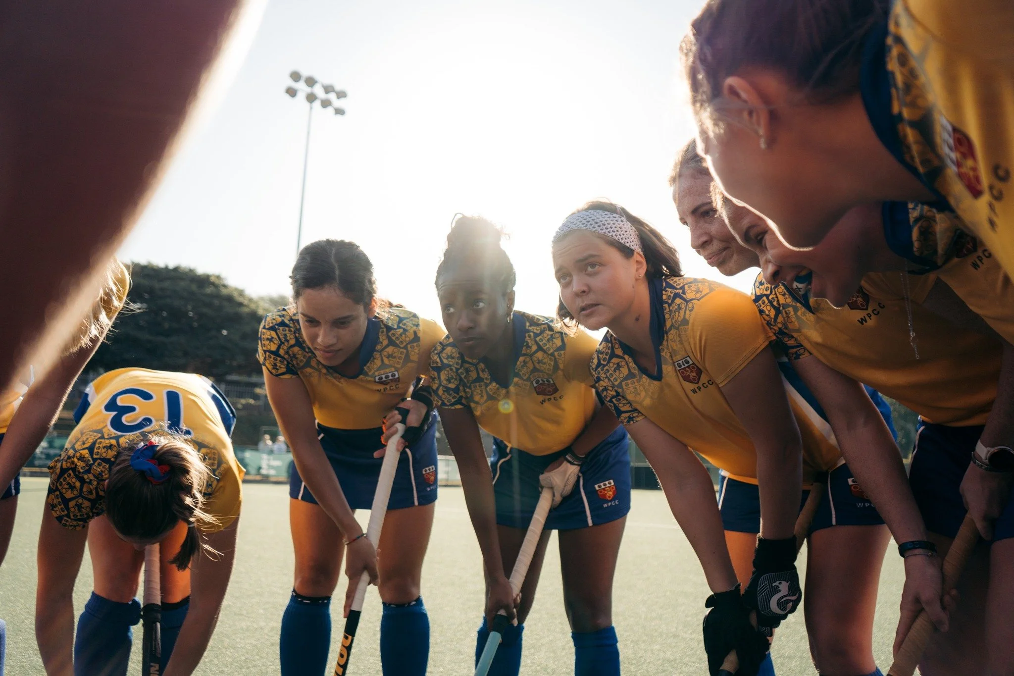 A group of female field hockey players in yellow and blue uniforms huddled together on the field during a game, with some players using sticks while listening attentively.