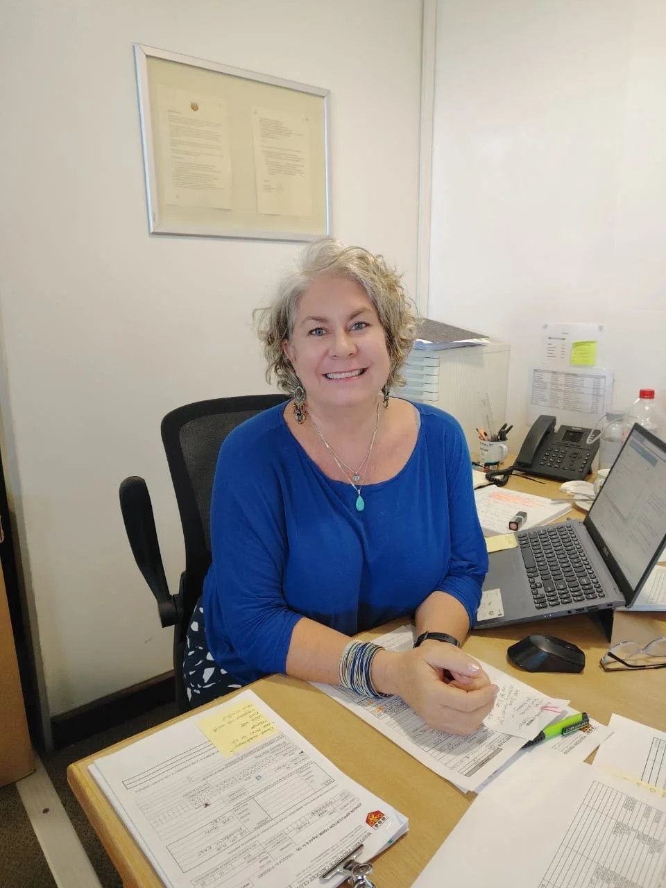 A woman with curly gray hair sitting at a cluttered office desk, smiling at the camera, wearing a blue shirt and jewelry.