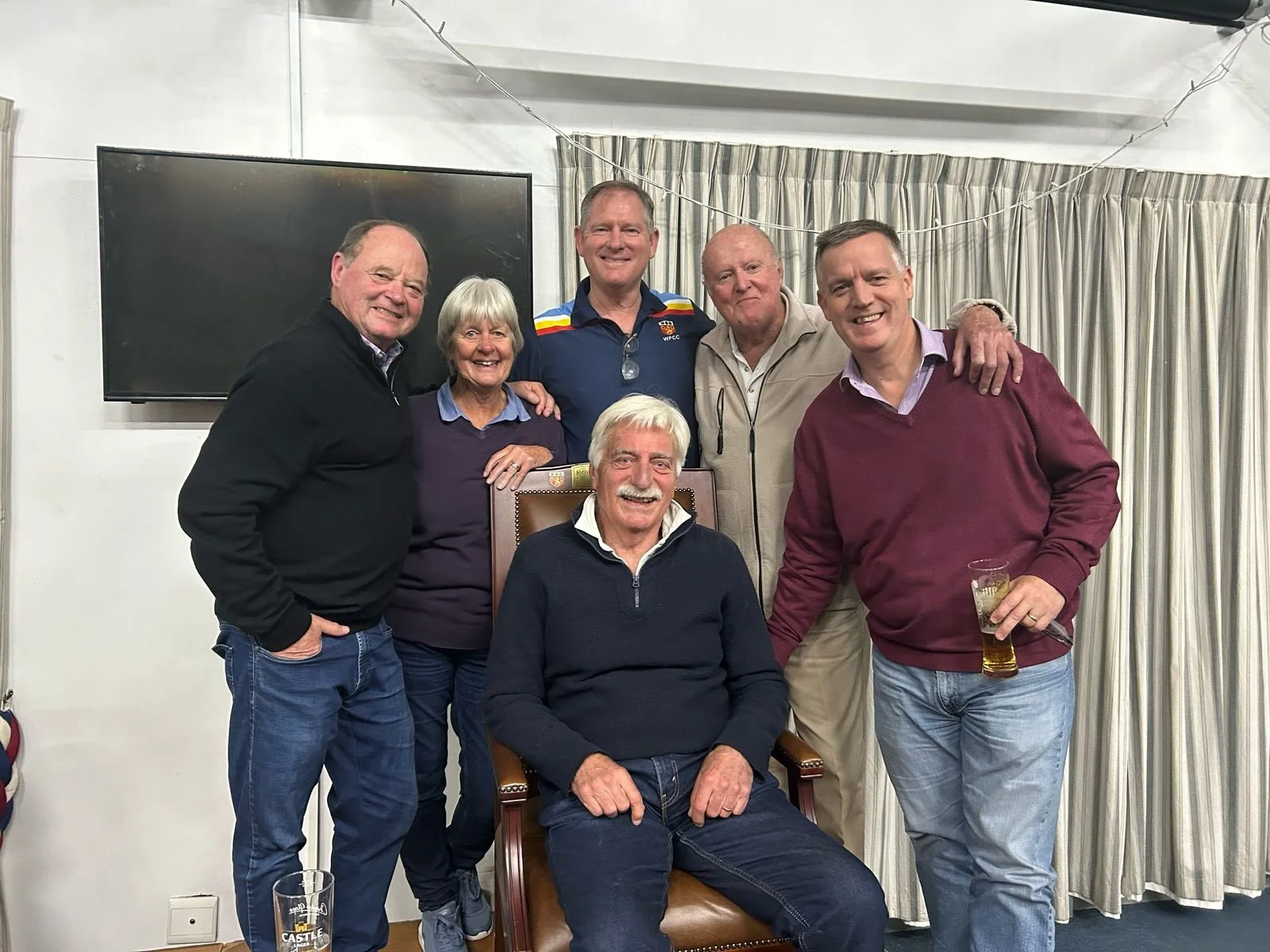 Group of six people, five men and one woman, smiling and posing for a photo indoors, with a man seated in a chair in front of them. A large flat-screen TV and curtains are visible in the background.