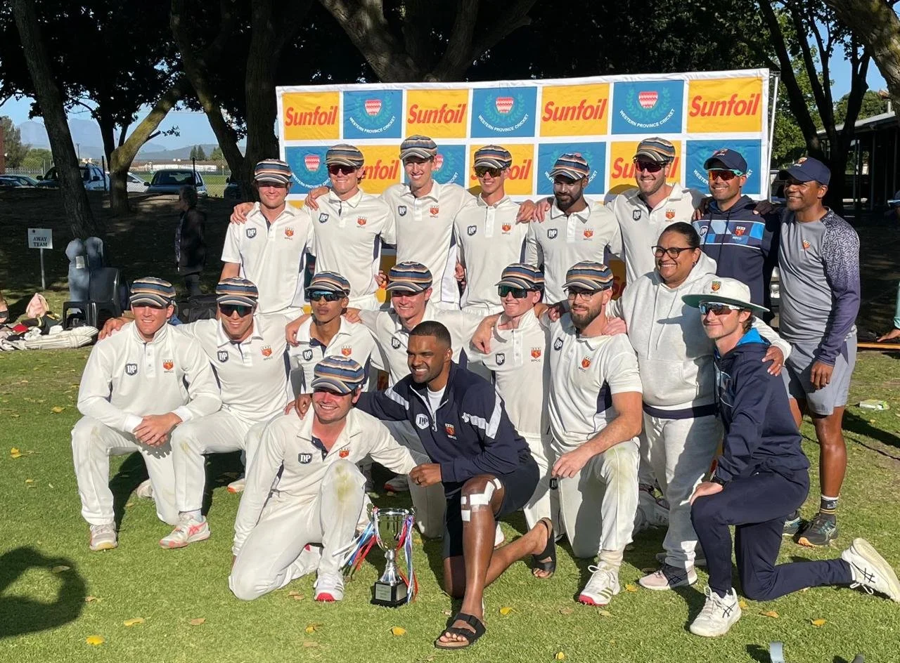 A group of cricket players posing with a trophy for a photo, outdoors on a sunny day, with trees and a banner in the background that says Sunfoil and features a logo. The players are dressed in white uniforms and caps, some are kneeling, others are standing, and one person is in sportswear.