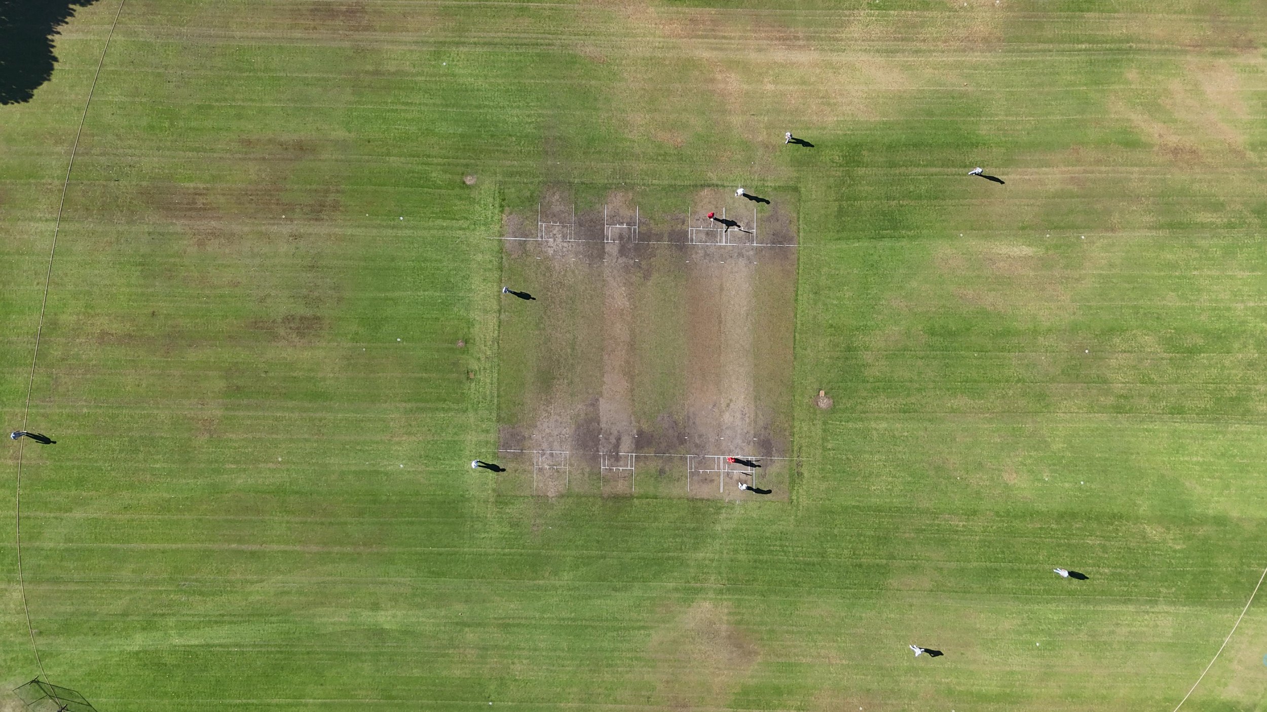 An aerial view of a grassy sports field with ten people standing around a central rectangular dirt area. Some people are wearing helmets, and the field is divided with lines and surrounded by a fence.