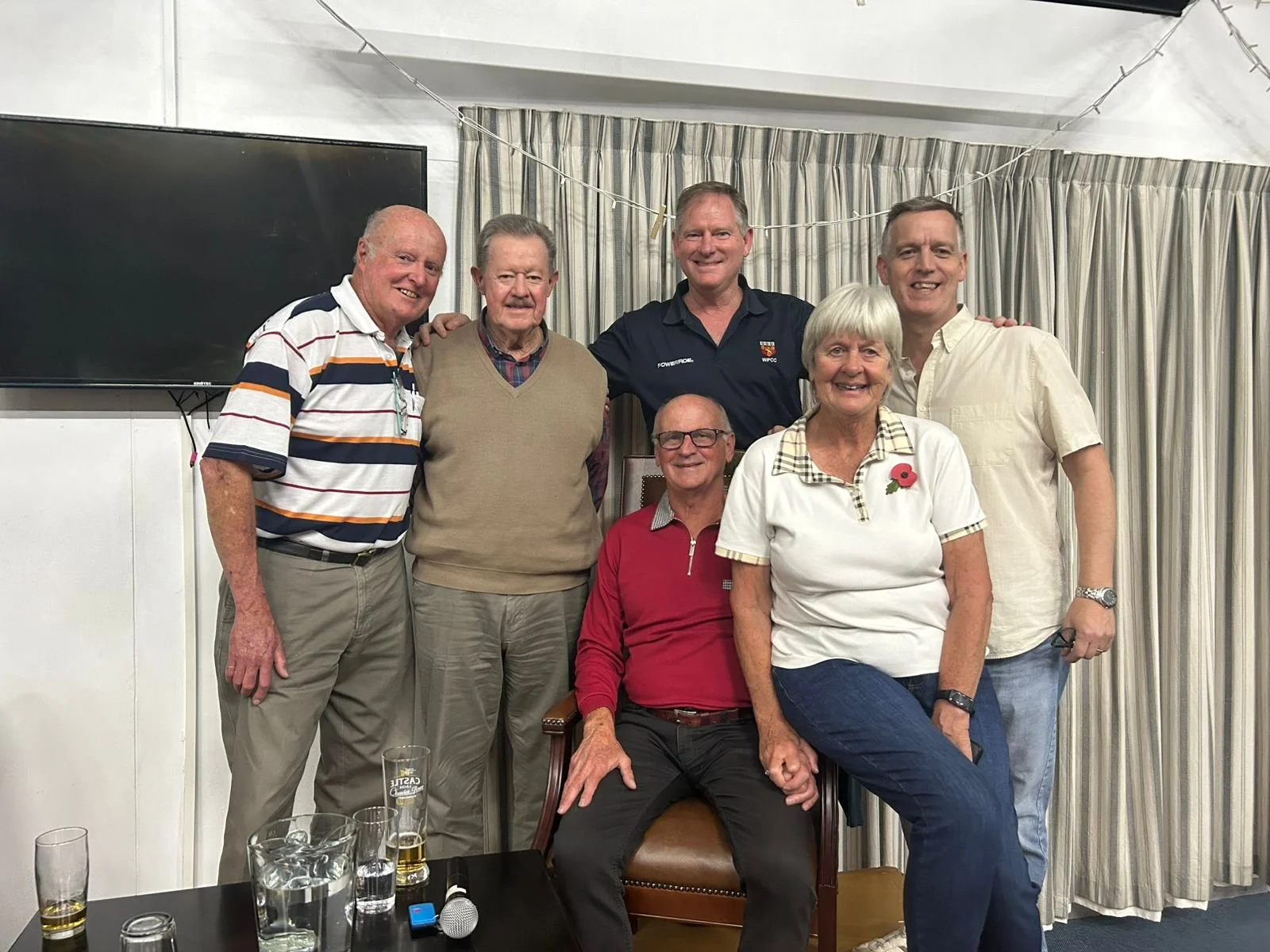Group of seven people, six men and one woman, posing together indoors, with drinks on a table in front of them and a large TV on the wall behind. They are smiling and standing close to each other.