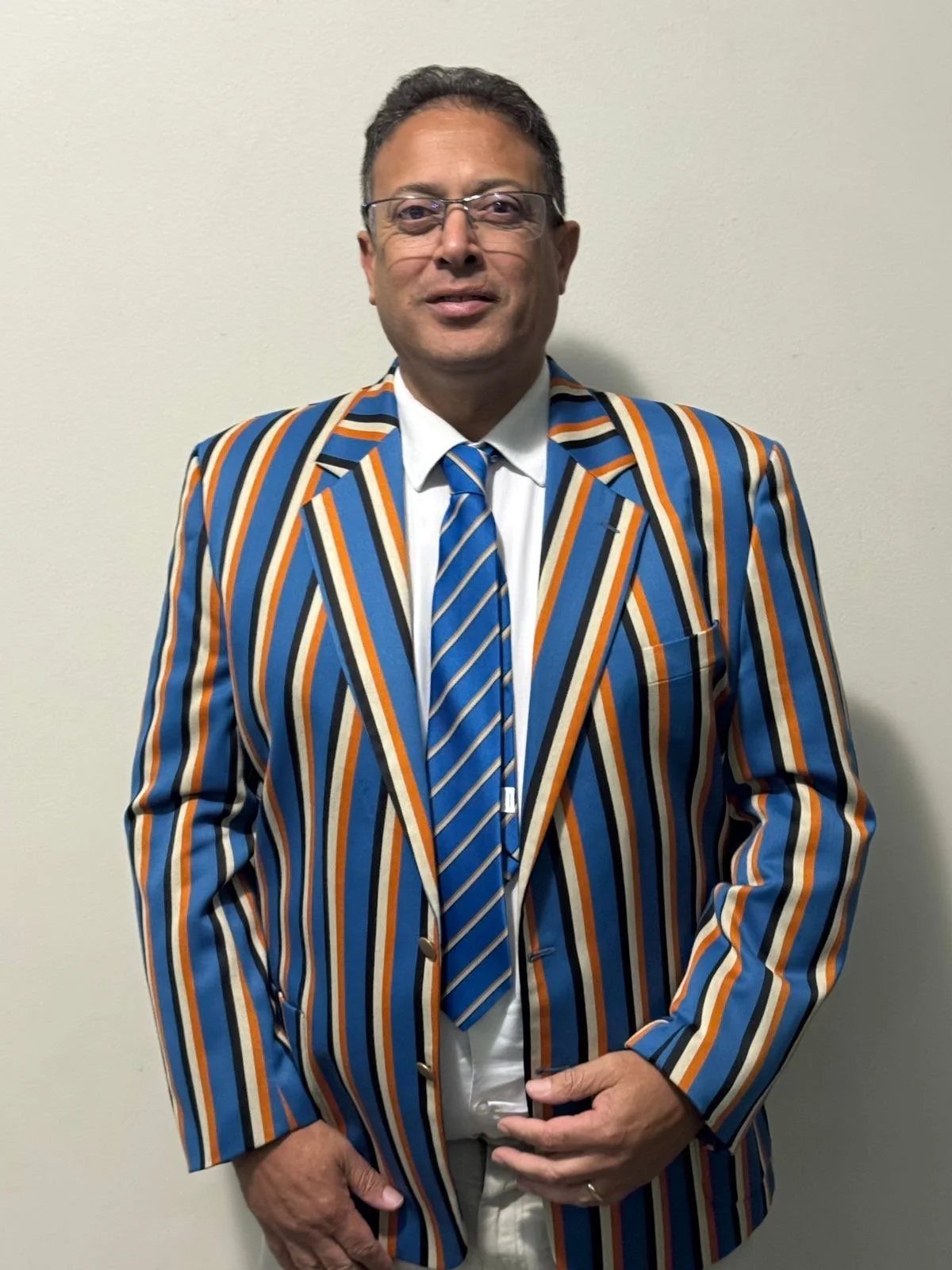 A man dressed in a multicolored striped suit with a white shirt and striped tie, standing against a plain light-colored wall.