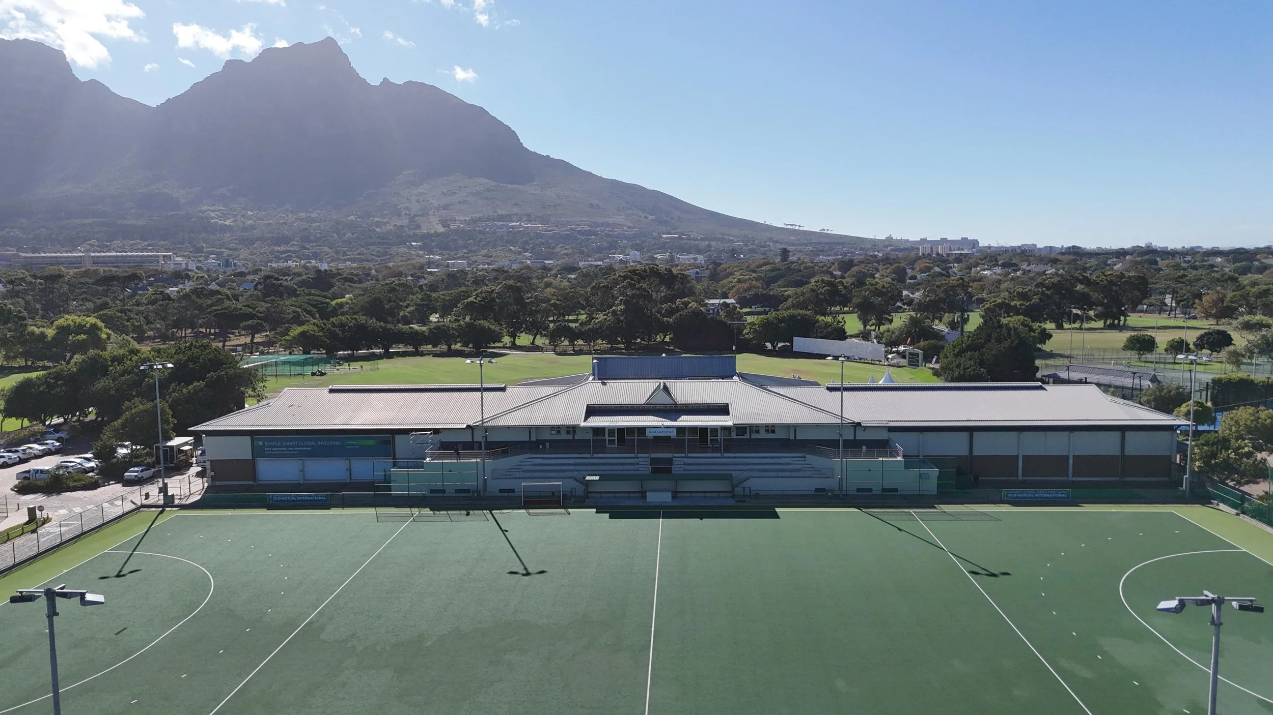 An outdoor sports field with a building behind it, surrounded by trees and mountains in the background, under a clear blue sky.