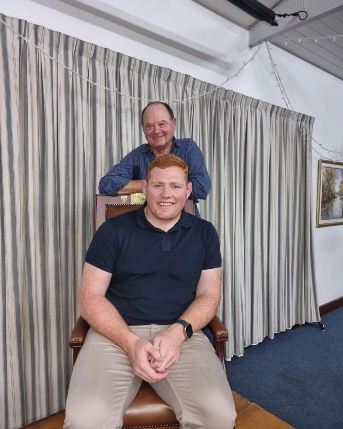Two men, one older and one younger, sitting in front of beige curtains and a ceiling with string lights. The older man is standing behind the younger man, resting his arms on the chair, both smiling at the camera.