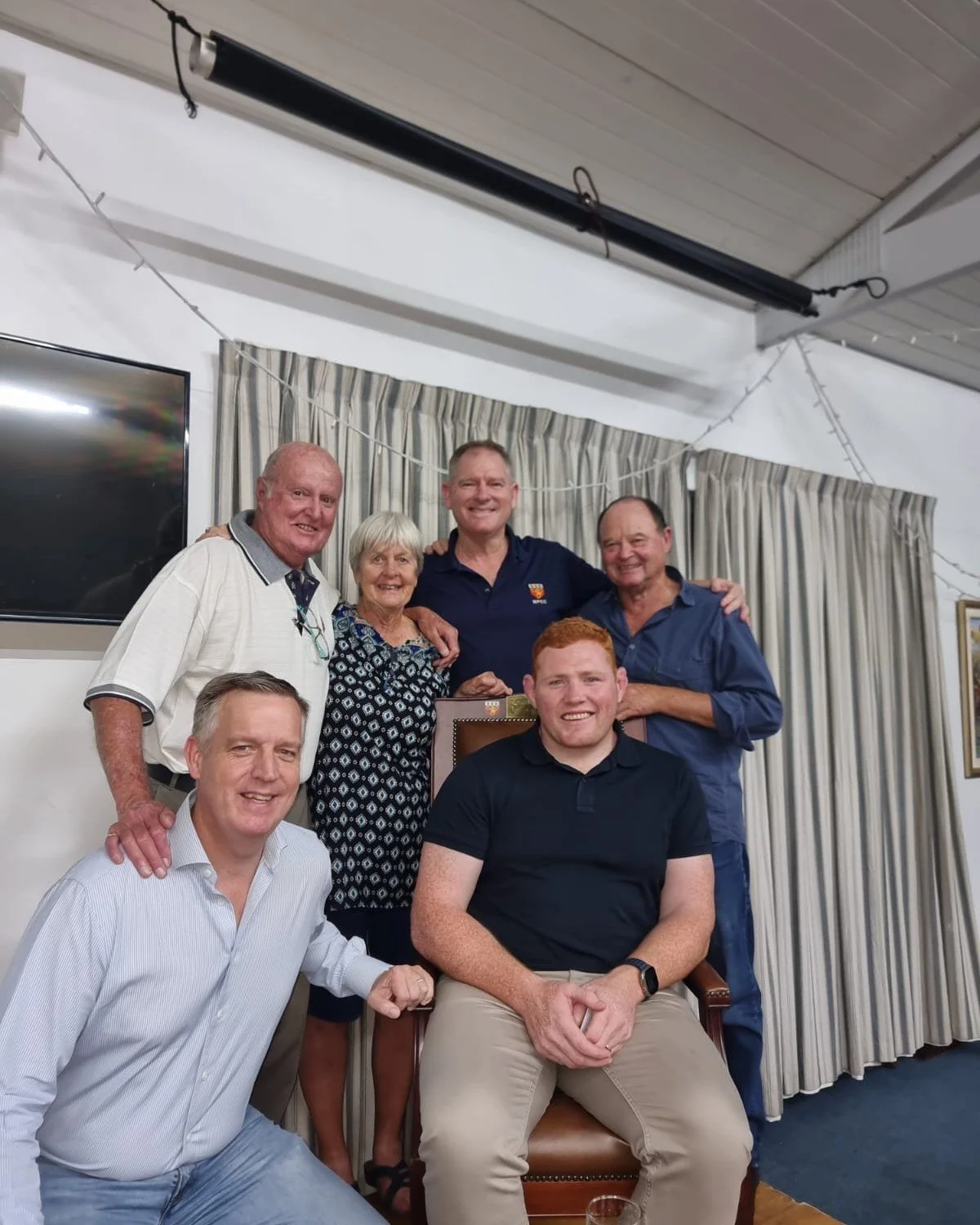 Group of seven smiling people gathered in a room with curtains and a TV, posing for a photo.