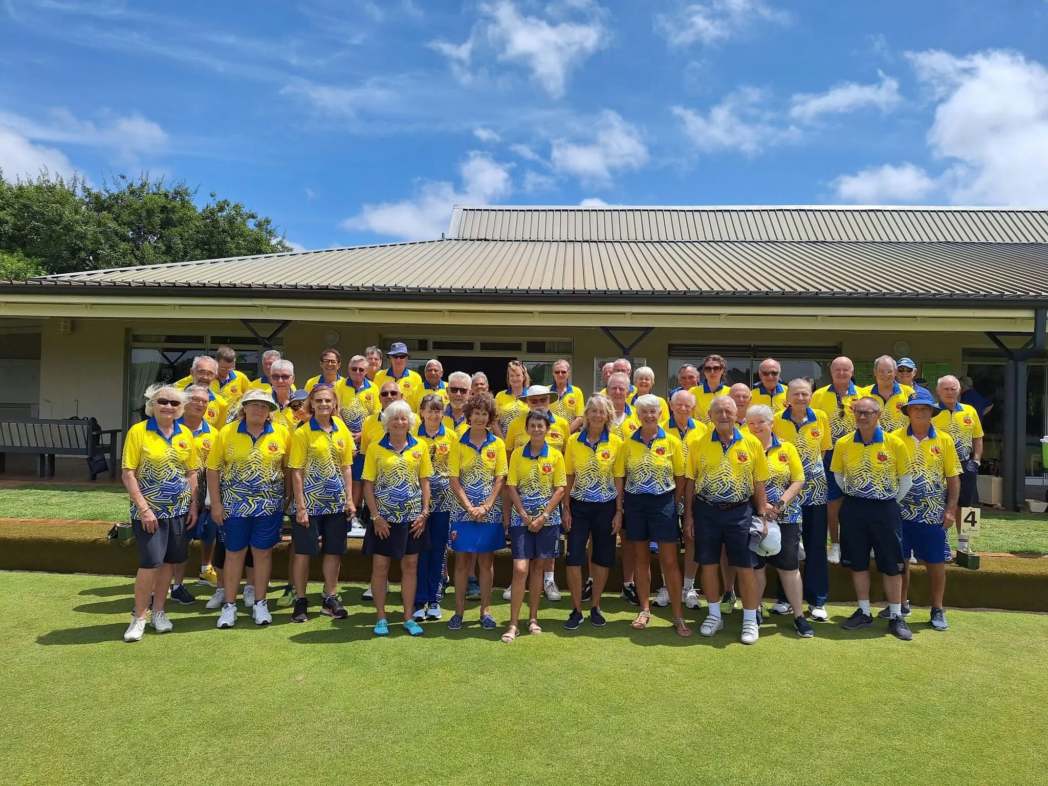 Group of people in yellow and blue sports shirts on a golf course with a clubhouse in the background under a blue sky.