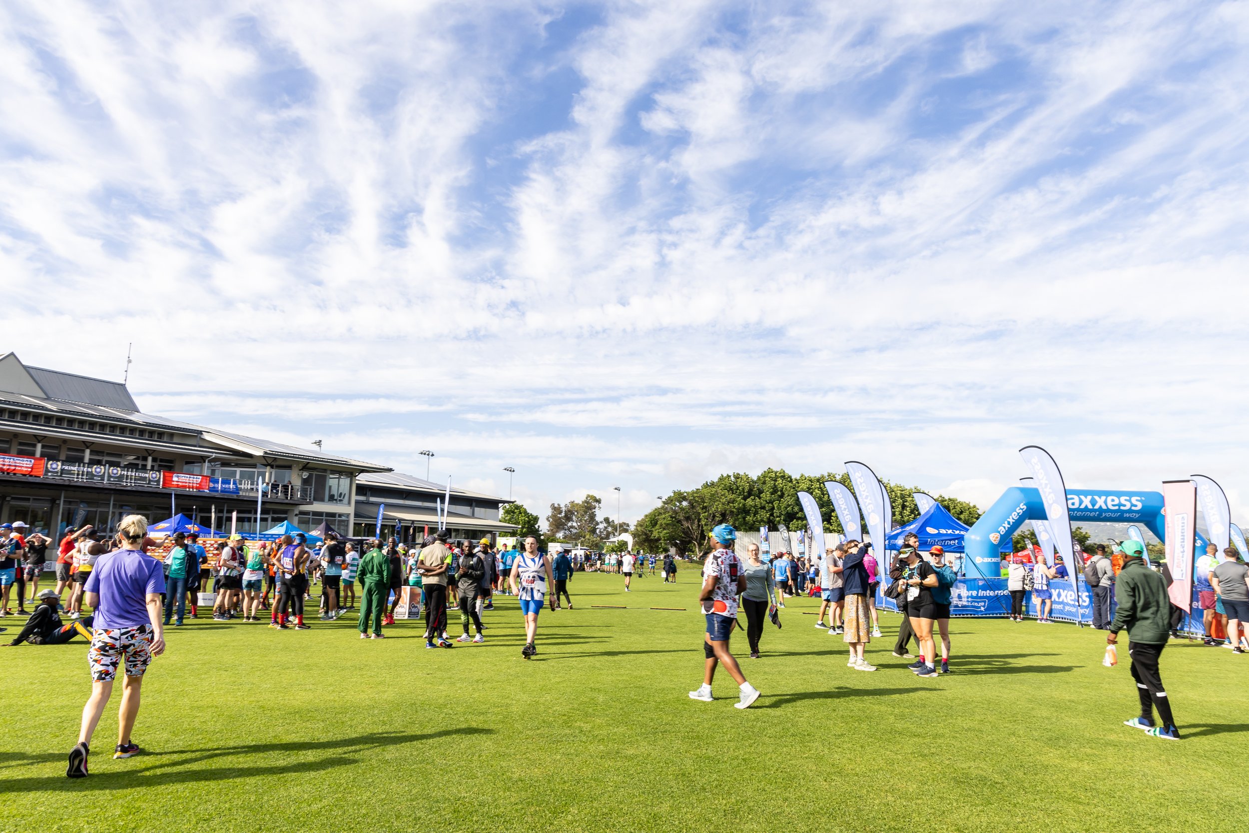 People gathered on a grassy field at an outdoor event, with tents and banners, and a building in the background under a partly cloudy sky.