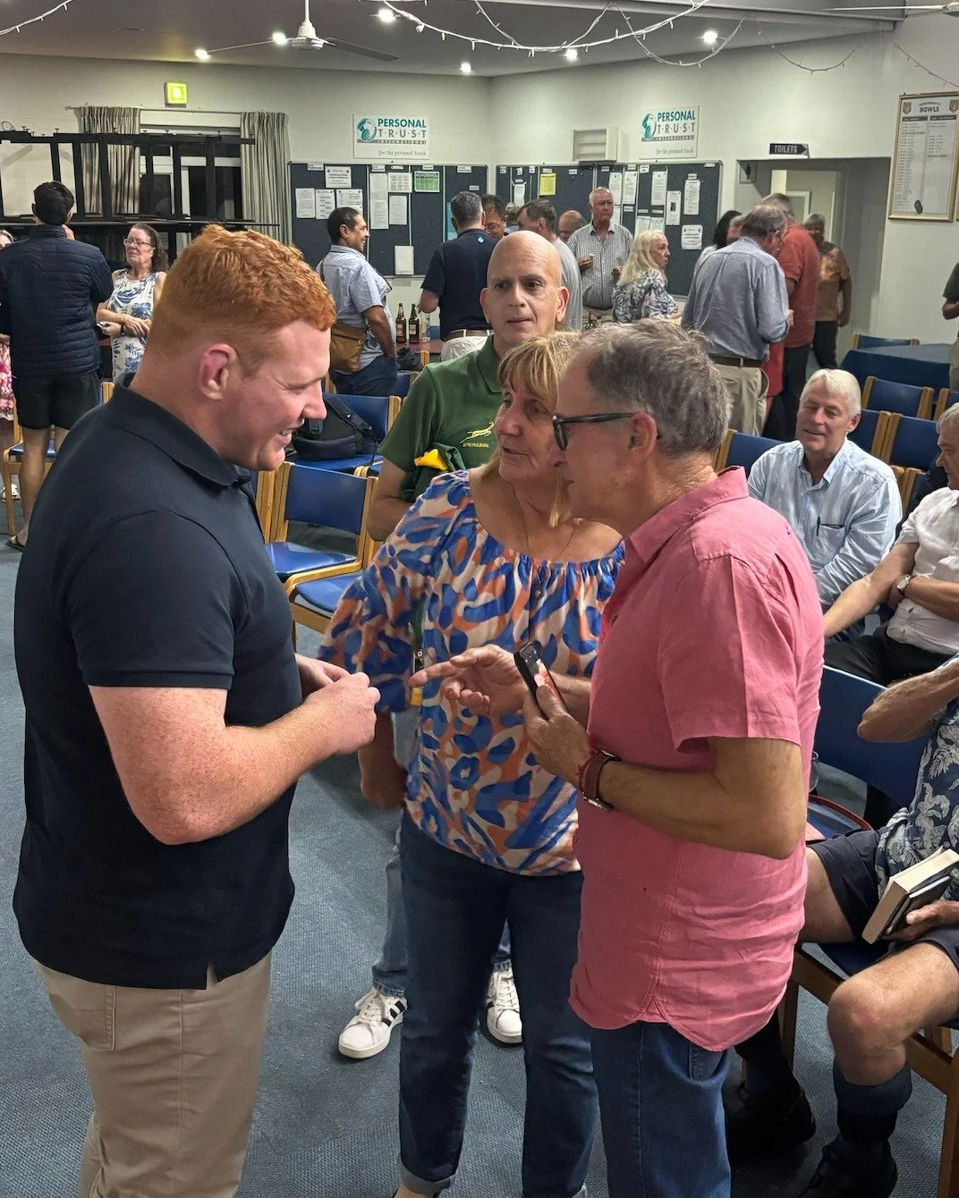 Group of people in a community hall engaging in conversation, some sitting and some standing, with bulletin boards and signs in the background.