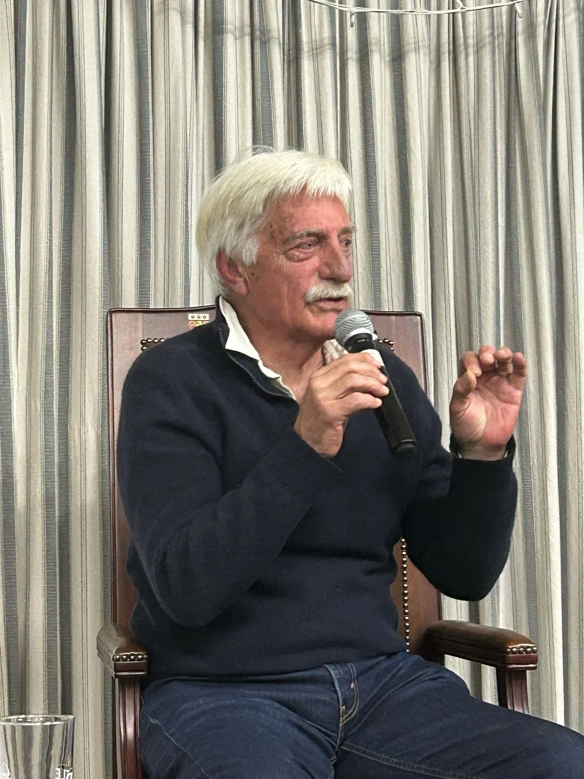 An elderly man with white hair and mustache speaking into a microphone while sitting in a brown chair against a backdrop of curtains.