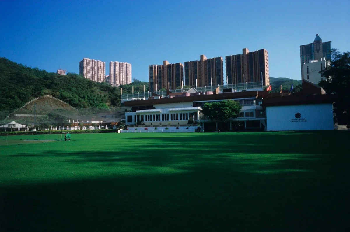 A cricket field with a clubhouse and high-rise buildings in the background, set against green hills under a clear blue sky.