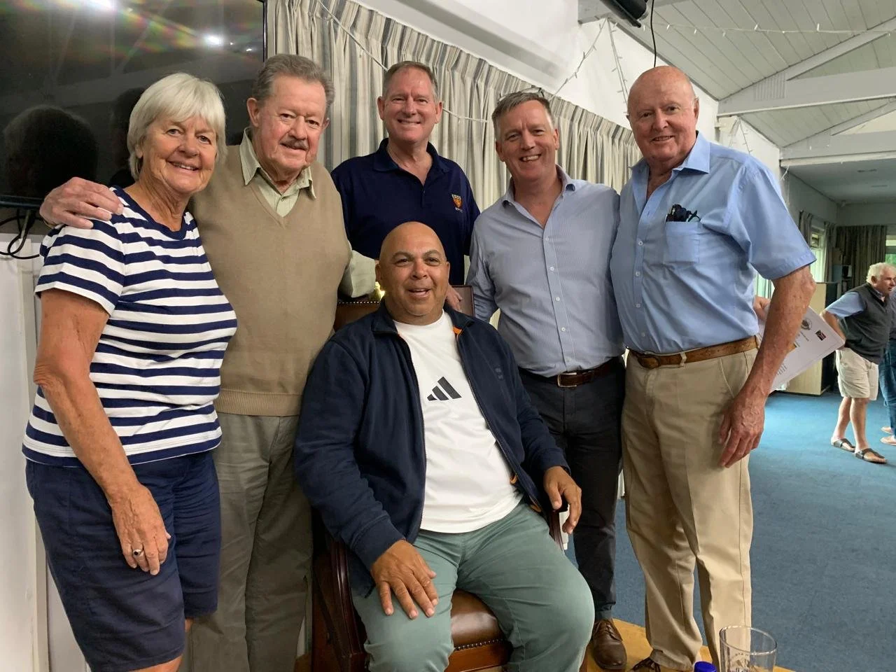 Group of six people smiling and posing together indoors, with some wearing casual clothes like shirts and sweaters, in a room with curtains and string lights.