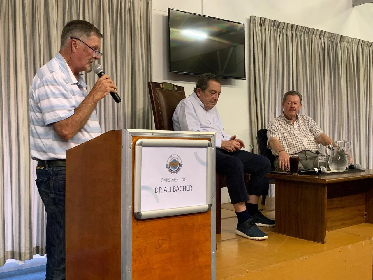 An older man with glasses and a striped shirt is speaking into a microphone at a podium labeled 'Dr. Ali Bacher' during a meeting. Two other men are seated on a stage behind him, one looking at his phone and the other sitting with arms on the table. 