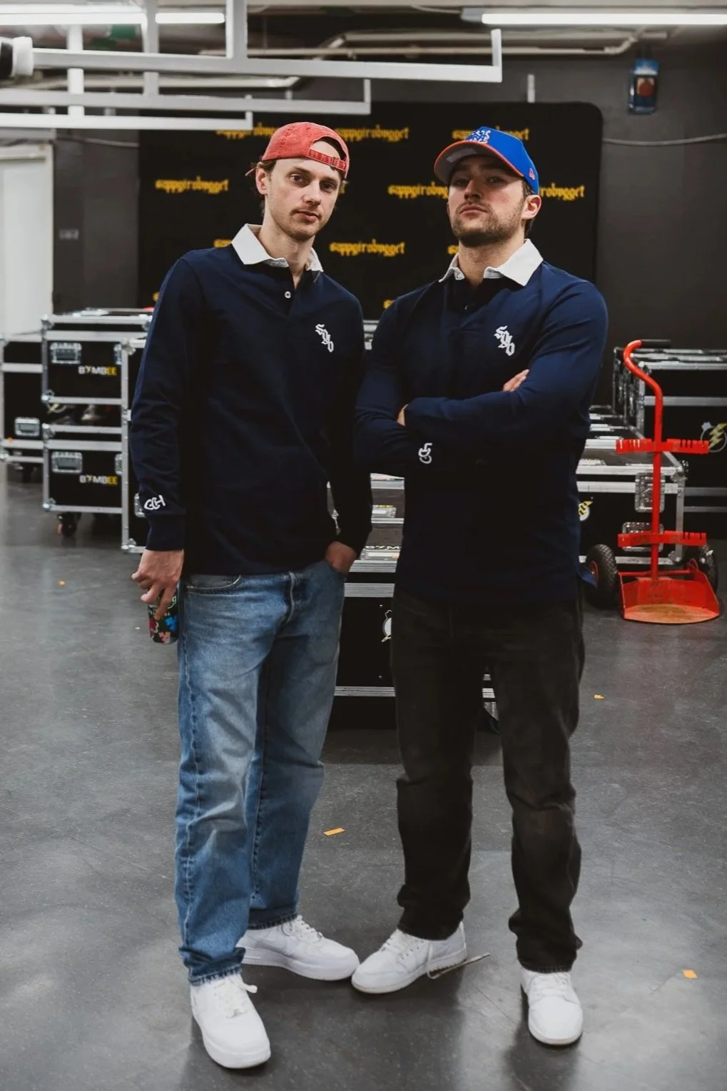 To unge menn står i en innendørs arena, begge har på seg like lyse jakker med logo. Den ene har rødt hette og lyse jeans, den andre har blå caps og svarte bukser. Bak dem er det utstyr og et rullende utstyrsstativ.