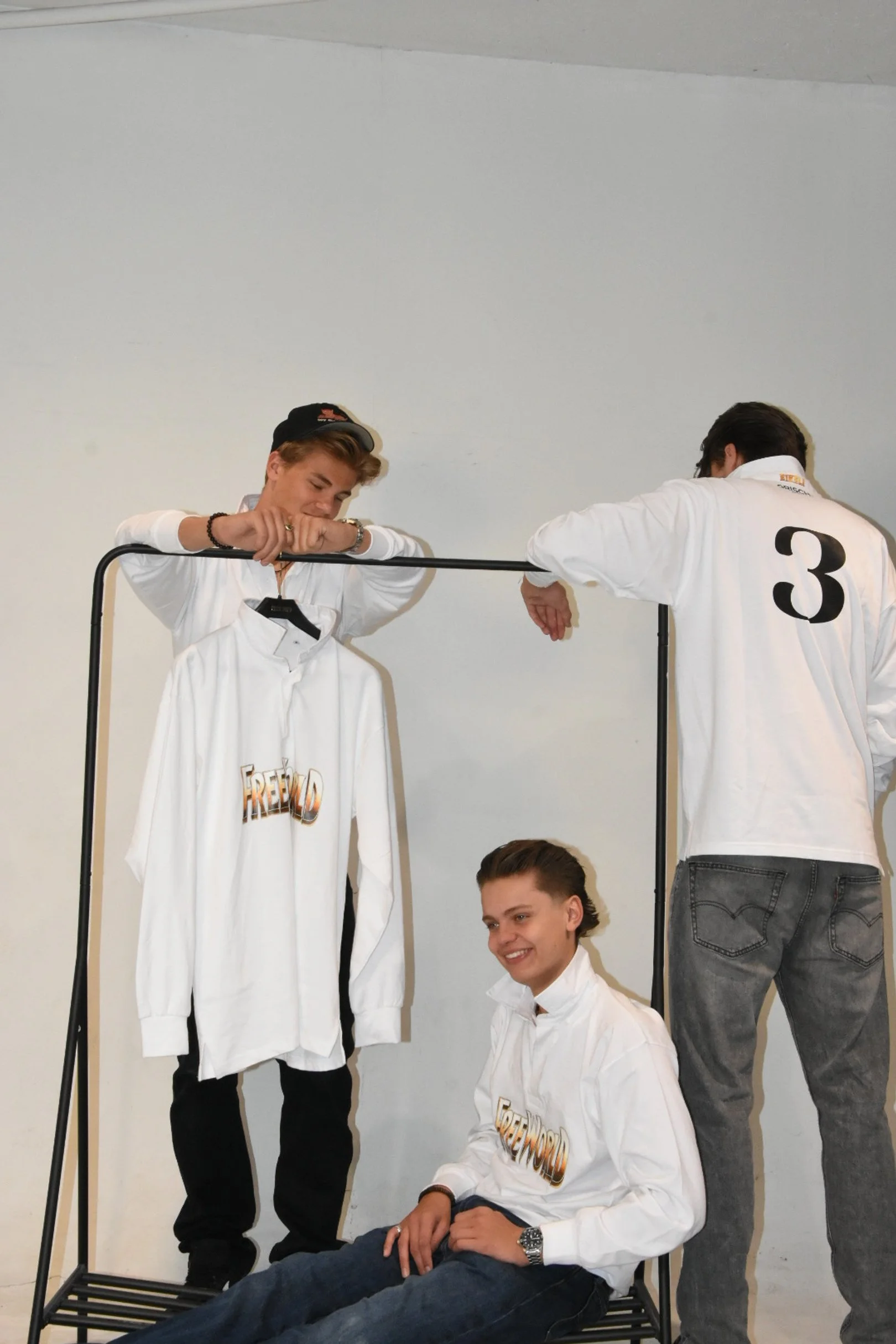 Tre unge menn som poserer sammen med et klesstativ med en hvit sweatshirt med teksten "FREEDOM" på, mot en hvit vegg.