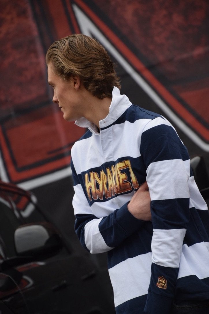 Person med langt, lyst hår i profil, iført en stripeet sportskjorte med logo som sier 'Hawkeye', står foran en bakgrunn med en rød og svart grafikk.
