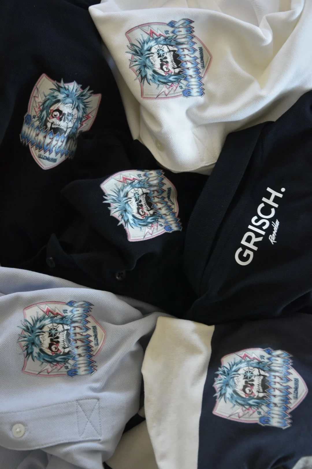 Kollektion av t-skjorter och en mössa med en design av en skrattande häxa med blå hår och ett korsade ben, samt en svart väska med texten 'GRISCH'.