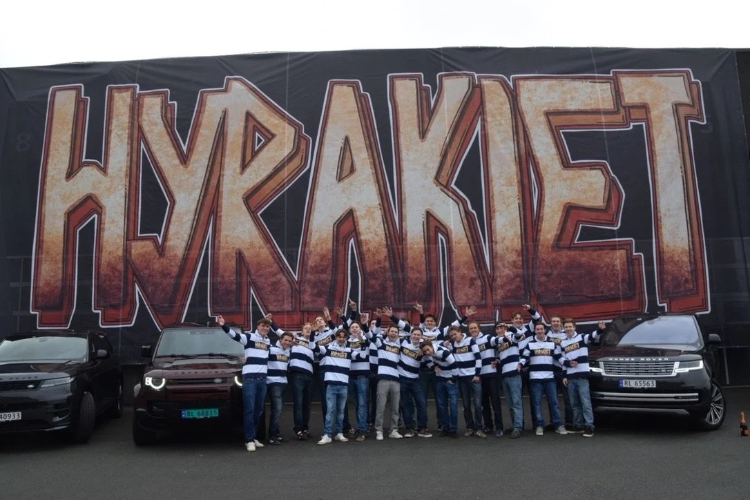 En gruppe mennesker i buret med mørke og hvite stripete drakter står foran en stor vegg med teksten "Hyraken" i graffiti-stil.