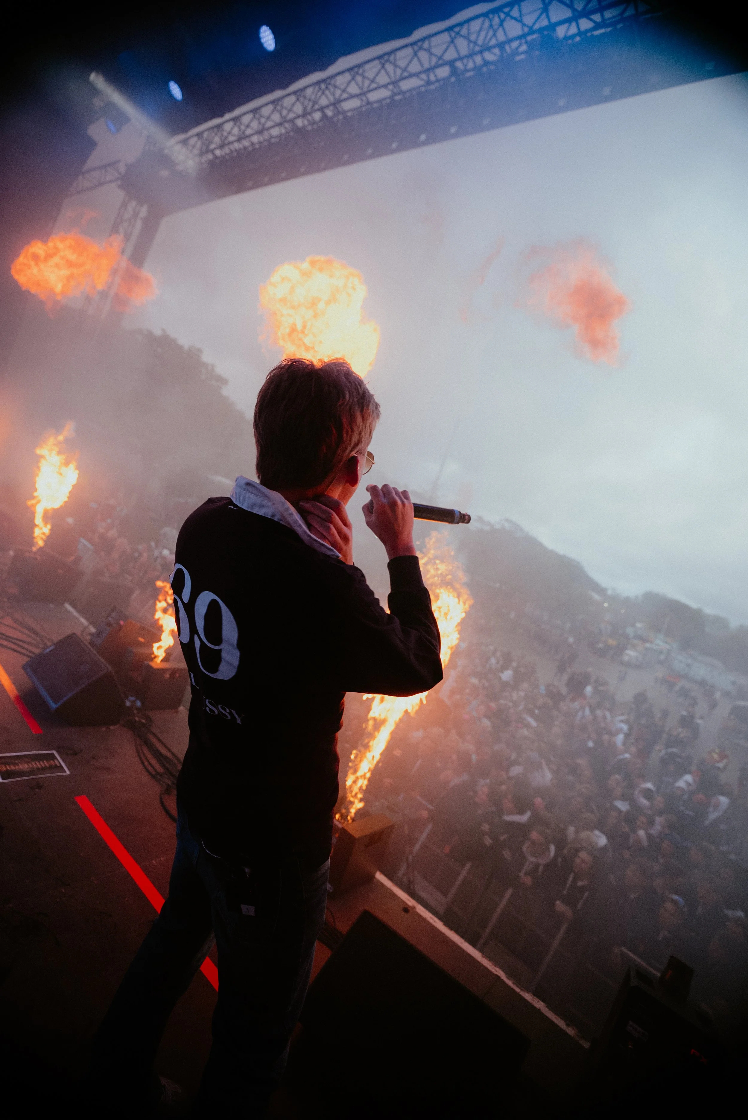 En person med mikrofon på en konsertscene, med ildfakler og en stor skjerm i bakgrunnen, og en stor publikum på utsiden.